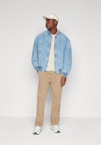 Hellblaues Jeans-Bomberjackett, beiges T-Shirt, beige Cargo-Hosen und weiße Sneaker. Komplett mit einer cremefarbenen Mütze und minimalen Details.