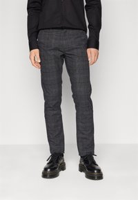 Pantalon à carreaux gris avec une coupe ajustée, confectionné dans un tissu texturé. Associé à des chaussures à lacets noires, offrant un look à la fois décontracté et raffiné.