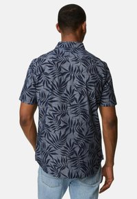 Mann in einem kurzärmligen, dunkelblauen Hemd mit einem dunklen Blattmuster, kombiniert mit hellblauen Jeans, seitlich gewandt.