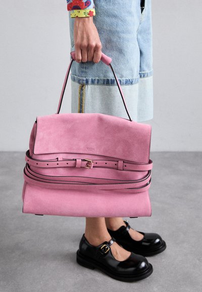 Borsa a mano in suede rosa con design strutturato, caratterizzata da una patta piegata, dettagli neri e un manico sottile. Indossata con pantaloni shorts di jeans e scarpe nere.