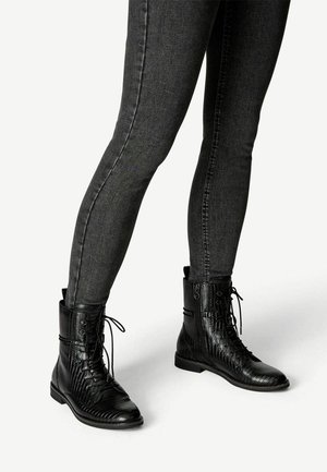 Botas de combate de cuero negro con patrón de cocodrilo texturizado, con cordones en la parte delantera y cremallera lateral, combinadas con jeans ajustados en gris oscuro.