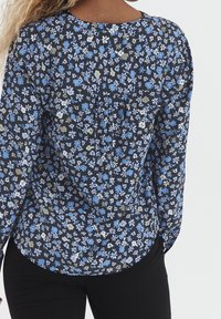 Blommig blus med svart bas, prydd med blå och vita blommor, långärmad, rundad halsringning och plisserad detalj på baksidan.