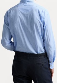 Mies, der ein hellblaues, gemustertes Langarmhemd trägt, das in dunkle marineblaue Anzughosen gesteckt ist, von hinten betrachtet.