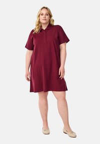Bordeaux polokleed met korte mouwen, knoopsluiting, A-lijn vorm en lengte tot op de knie. De stof lijkt zacht met een gladde textuur.