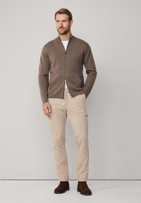 Muž oblečený v hnedšom kardigane na zips, béžových cargo nohaviciach a tmavohnedých čižmách, stojí s jednou rukou vo vrecku proti jednoduchej pozadia.