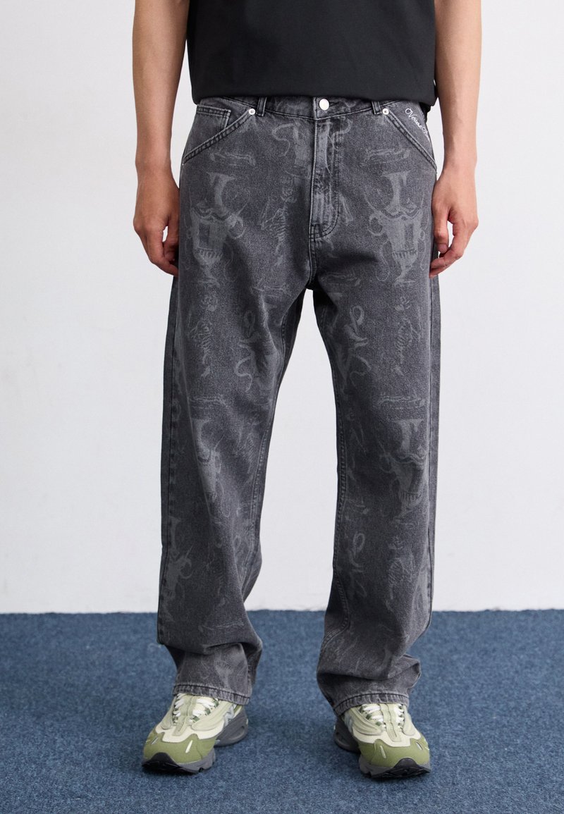 Jeans de mezclilla gris oscuro con un patrón texturizado, corte holgado, cinco bolsillos y detalles bordados, combinados con zapatillas acentuadas en verde.