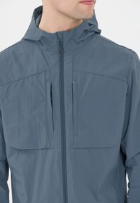 Lätt blå jacka med huva, med en framzipper, två vertikala bröstfickor och en slät yta. Hållbart och väderbeständigt material.