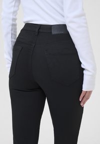 Jeans en denim noir à taille haute, avec un design à cinq poches et des coutures contrastantes. La ceinture comporte une patch en cuir avec une marque.