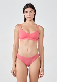 Donna in piedi di fronte, che indossa un reggiseno e un perizoma coordinati color corallo contro uno sfondo chiaro e semplice.
