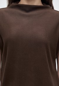 Gros plan d'une personne portant un haut marron simple avec un col montant légèrement plié et une texture de tissu lisse.