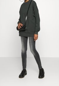 Tmavě zelená oversized mikina s nízkými rameny, doplněná šedými slim-fit džínami a černými polobotkami na tkaničky. Černá crossbody taška s kruhovými detaily.