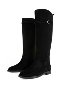 Bottes en suède noir montantes, avec un bout rond, un talon plat et une sangle décorative avec des accessoires en or près du haut. Design simple et élégant.