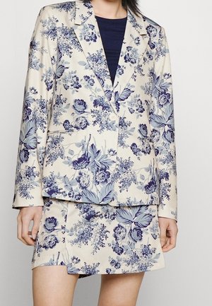 Kvinde iført et sæt med cremefarvet blazer og nederdel med blå blomsterprint, kombineret med en mørkeblå top under.