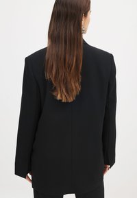 Must mustritud must blazer, millel on kohandatud õlad, sile tekstuur ja pikk seljaosa disain. Omab minimalistikku aktsentide kogumit ja puhast siluetti.