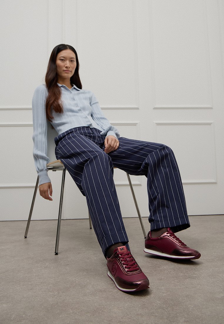 Donna con lunghi capelli scuri, che indossa una camicia azzurro chiaro, pantaloni a righe navy e sneakers borgogna, seduta in modo rilassato su una sedia di metallo all'interno.