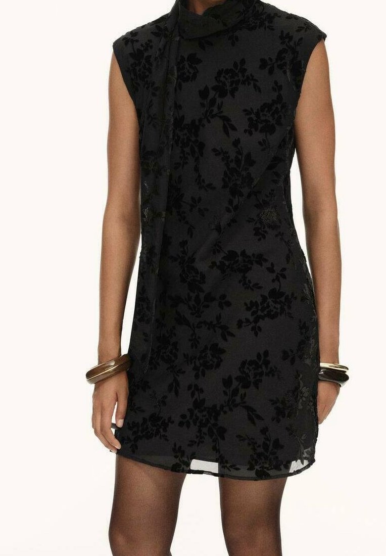 Robe noire sans manches avec motif floral en velours, col haut avec un nœud sur le côté, portée avec des collants transparents foncés et des bracelets métalliques aux deux poignets.