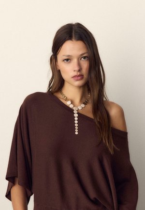 Mlada žena s dugom smeđom kosom nosi smeđu bluzu s otvorenim ramenima i zlatno-bijelu ogrlicu s privjeskom u obliku kruga na jednostavnoj svijetloj pozadini.