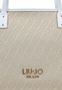 Beige tygkasse med ett vävt mönster, med vita handtag i konstläder och en guldfärgad logotyp "LIU·JO MILANO" i mitten.