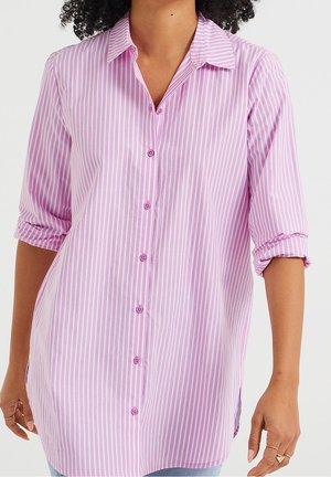 Kvinde iført lang pink og hvid vertikalt striped skjorte med opkørte ærmer og lyseblå jeans.