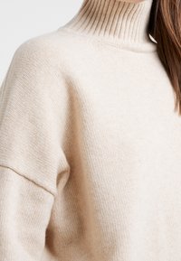 Pull en tricot beige avec un col à côtes haut, texture douce et épaules tombantes. Le tissu présente un motif fin et uniforme.