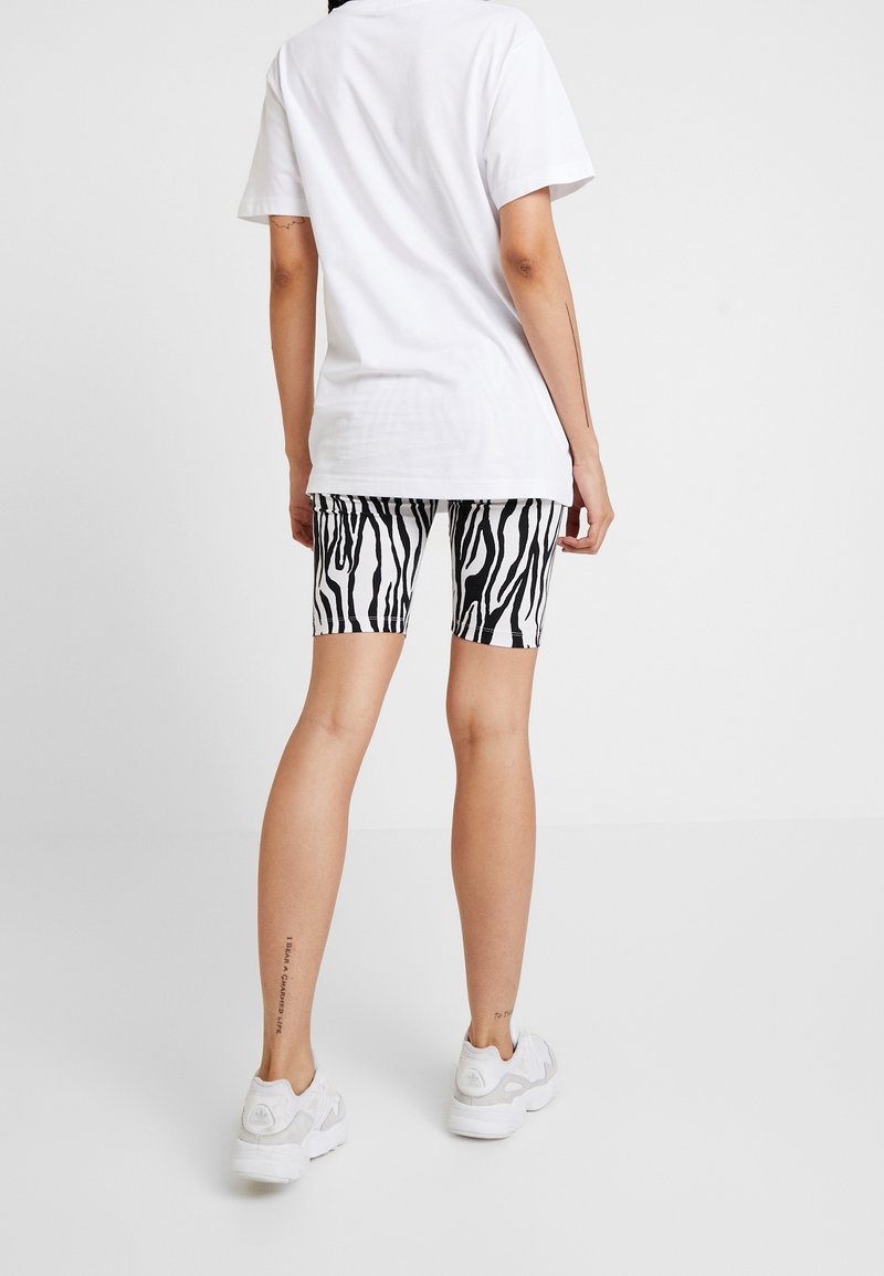 Černobílé kraťasy se zebrovaným potiskem spárované s jednoduchým bílým tričkem a světle šedými teniskami. Viditelné tetování na noze poblíž kotníku.