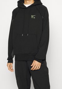 Person som bär en svart Nike Air-hoodie med en framficka och svarta Nike-byxor med en synlig logotyp på vänster lår.