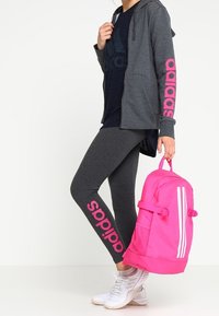Grijze rits-hoodie met roze "adidas" tekst op de mouwen, gecombineerd met grijze legging die hetzelfde logo heeft. Felroze rugtas met witte strepen.