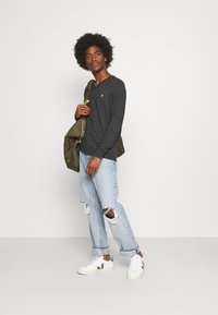Camisa gris de manga larga con cuello en V, logo amarillo en el pecho, combinada con jeans claros desgastados y zapatillas blancas. Bolsa verde oliva.
