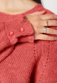 Gebreide trui in zacht koraal met een fuzzy textuur. Bevat twee glanzende roze knopen bij de manchetten. Hand versierd met ringen.