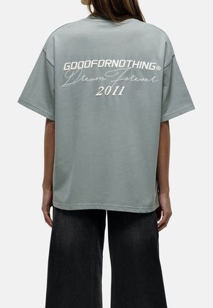 Femme vue de dos portant un T-shirt gris ample avec "GOODFORNOTHING Dream Forever 2011" imprimé dessus et un pantalon large noir.