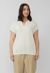Kobieta ubrana w beżowe spodnie o szerokich nogawkach oraz białą bluzkę w żółte paski z krótkim rękawem i dekoltem w serek, stojąca na jednolitym tle.