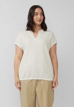 Vrouw in beige wijde broek en een kortemouwen witte en lichtgele gestreepte V-hals top, staand tegen een effen achtergrond.