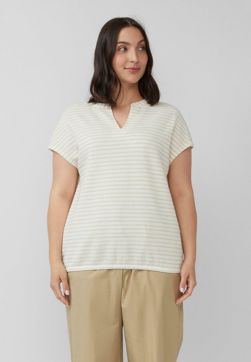 Kobieta ubrana w beżowe spodnie o szerokich nogawkach oraz białą bluzkę w żółte paski z krótkim rękawem i dekoltem w serek, stojąca na jednolitym tle.