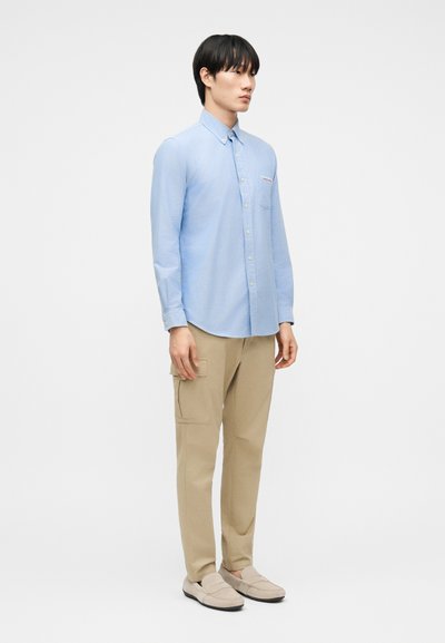 Νέος άνδρας που στέκεται φορώντας ανοιχτό μπλε πουκάμισο με κουμπιά, μπεζ παντελόνι καρό και μπεζ παπούτσια χωρίς κορδόνια, μπροστά σε απλό λευκό φόντο.