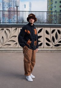 Svart jacka med orange och bruna mönster, beige cargobyxor, vita sneakers och en brun pälsmössa. Urban bakgrund med dekorativ vägg.