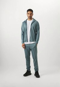 Atletska trenirka v svetlo modrozelene barve, narejena iz gladke, zračne tkanine. Sestavljena je iz jakne s kapuco na zadrgo in ujemajočih se hlač, obuta v črne superge.