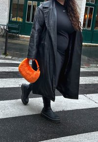 Svart läderjacka över svart outfit, bärande på en fluffig orange handväska. Tjocksålda svarta stövlar, som går över ett övergångsställe.