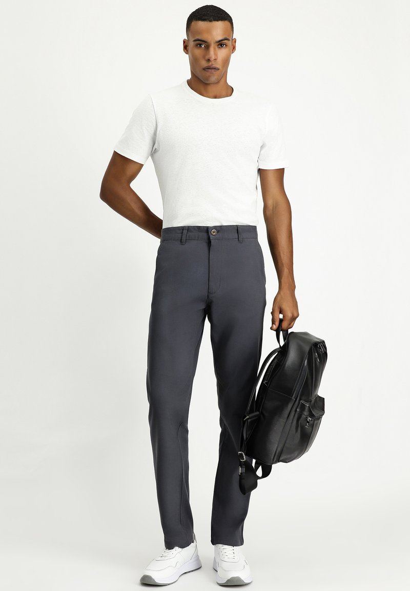 Pantalones grises con cierre de botón, combinados con una camiseta blanca lisa y una mochila negra, junto con zapatillas blancas. Textura de tela suave.