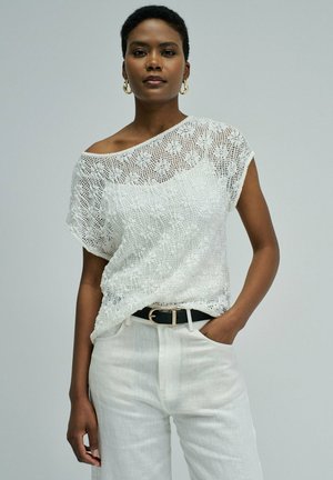 Mujer con cabello corto que lleva una blusa blanca de encaje y pantalones blancos, de pie con una mano en el bolsillo contra un fondo liso.