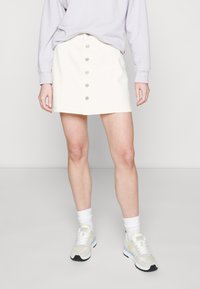 Jupe mini en denim blanc avec fermeture à boutons sur le devant et cinq boutons, associée à un sweat-shirt violet clair et des baskets blanches.