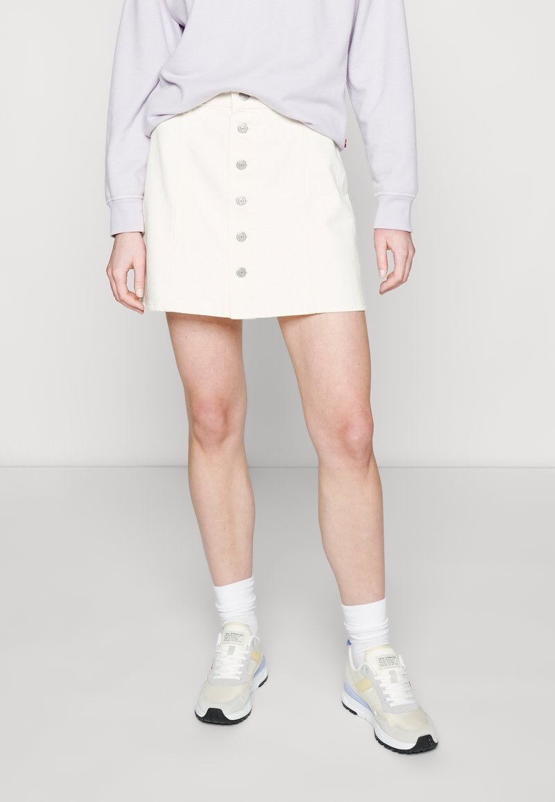 Jupe mini en denim blanc avec fermeture à boutons sur le devant et cinq boutons, associée à un sweat-shirt violet clair et des baskets blanches.