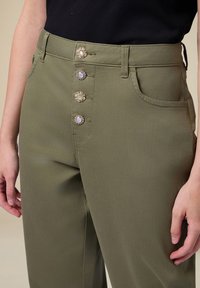 Olivgrüne hochtaillierte Hose mit glatter Textur, fünf dekorativen Knöpfen in Goldfarbe und Seitentaschen. Nahaufnahme.