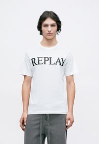 Mladenič s srednje dolgimi temnimi lasmi nosi belo majico s sloganom "REPLAY" in ohlapne sive hlače pred enostavno belo ozadje.