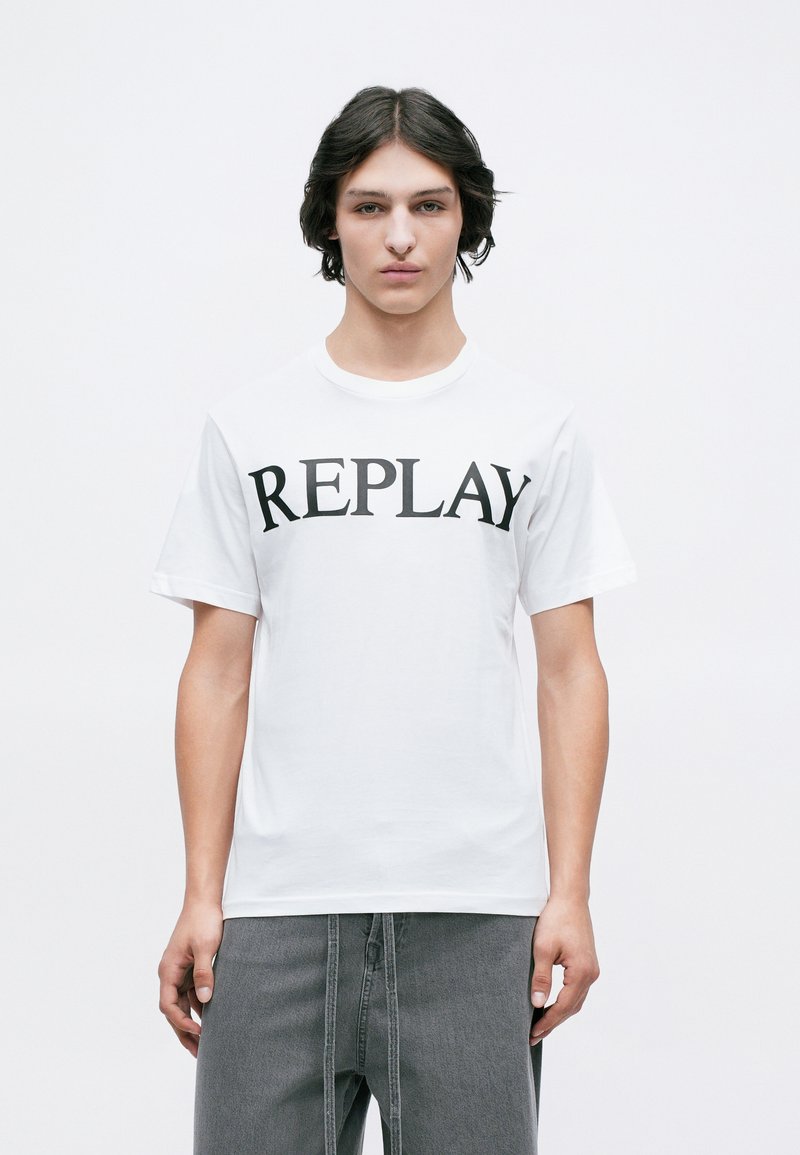 Mladenič s srednje dolgimi temnimi lasmi nosi belo majico s sloganom "REPLAY" in ohlapne sive hlače pred enostavno belo ozadje.