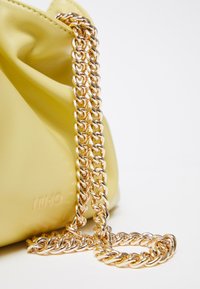 Sac en cuir jaune avec une épaisse chaîne dorée en bandoulière, posé sur une surface blanche, montrant une partie du nom de la marque embossé "LIU·JO".