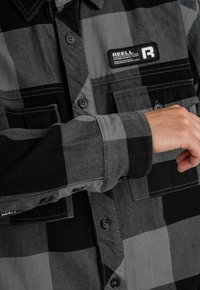 Schwarz-graues karierter Hemd mit einem Knopfverschluss-Design. Mit einem Markenpatch und zwei vorderen Taschen mit schwarzen Knöpfen. Aus Baumwollmaterial.