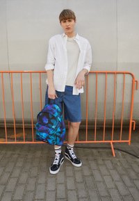 Plecak w niebieski kwiatowy wzór z czarnymi akcentami. Model nosi białą koszulę w paski, beżowy t-shirt, dżinsowe szorty oraz czarne trampki.