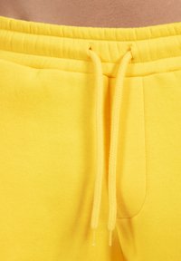 Żółte spodnie dresowe wykonane z miękkiego materiału, z elastycznym pasem i sznurkiem ściągającym oraz boczną kieszenią. Gładka tekstura, prosty design.