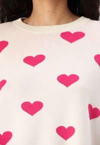 Pletena majica u krem boji s okruglim izrezom, ukrašena raširenim magenta srcima kao uzorkom, meke teksture i ležernog kroja.