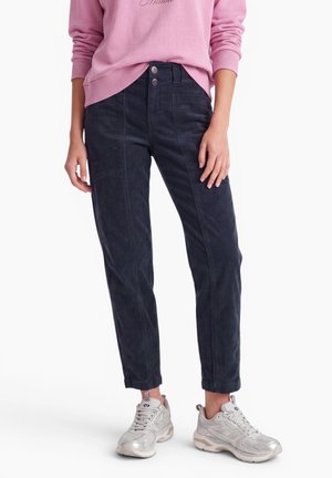 Personne portant un pantalon sombre fuselé avec des revers retroussés, un haut à manches longues rose et des baskets argentées, debout sur un fond blanc.
